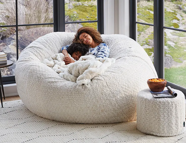 Woman and child relaxing on Lovesac Sac large bean bag chair with a blanket and Squattoman footstool in living room