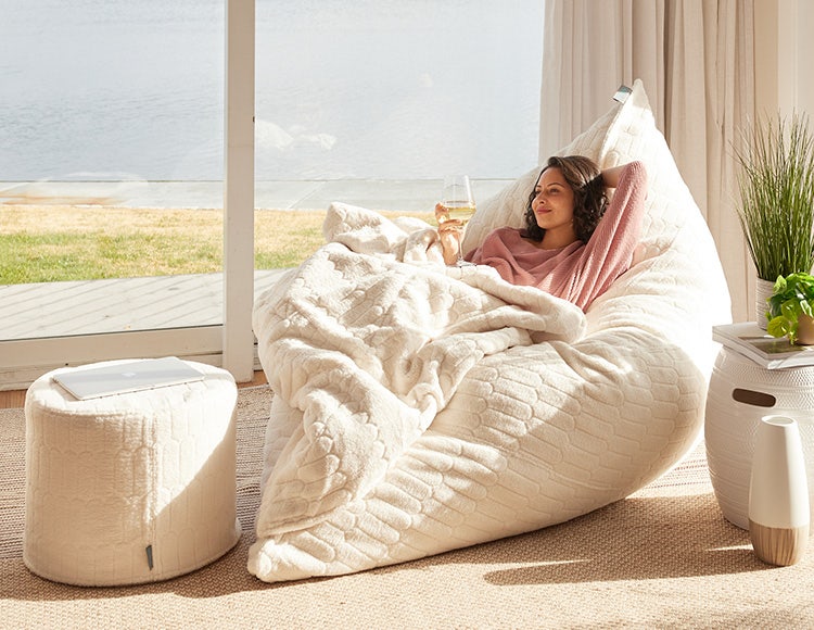 Woman relaxing on Lovesac Sac large bean bag chair with a blanket and Squattoman footstool